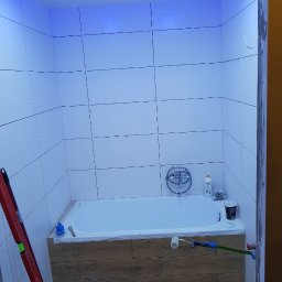 KA RENOVATION - Łazienka w trakcie remontu w Warszawie: białe płytki na ścianach, obudowana drewnopodobnymi panelami wanna, niebieskie oświetlenie sufitowe, poziomica oparta o ścianę, wałek malarski i akcesoria...