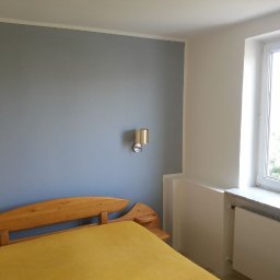 KA RENOVATION - Pomieszczenie po malowaniu ścian na szaro, z drewnianym łóżkiem, lampką nocną i białym oknem z widokiem na zieleń.