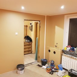 KA RENOVATION - Pokój w trakcie odświeżania: ściany pomalowane na beżowo, widoczne puszki z farbą, narzędzia malarskie i grzejnik pod oknem. W tle otwarte drzwi do innego pomieszczenia z regałem.