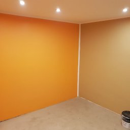 KA RENOVATION - Pomarańczowa i beżowa ściana w pokoju po świeżym pomalowaniu, widoczna puszka z farbą przy podłodze.