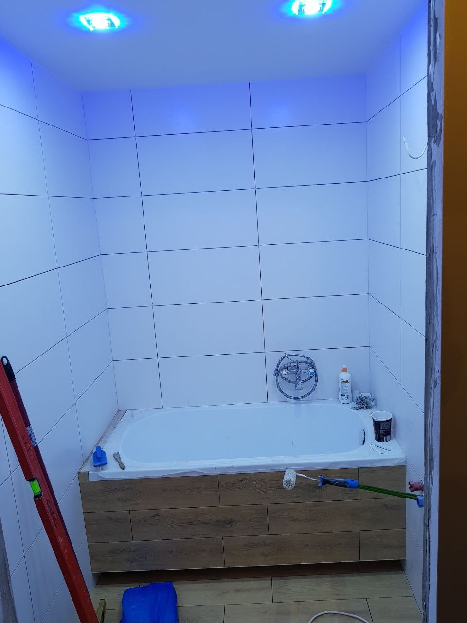 Łazienka w trakcie remontu w Warszawie: białe płytki na ścianach, obudowana drewnopodobnymi panelami wanna, niebieskie oświetlenie sufitowe, poziomica oparta o ścianę, wałek malarski i akcesoria...