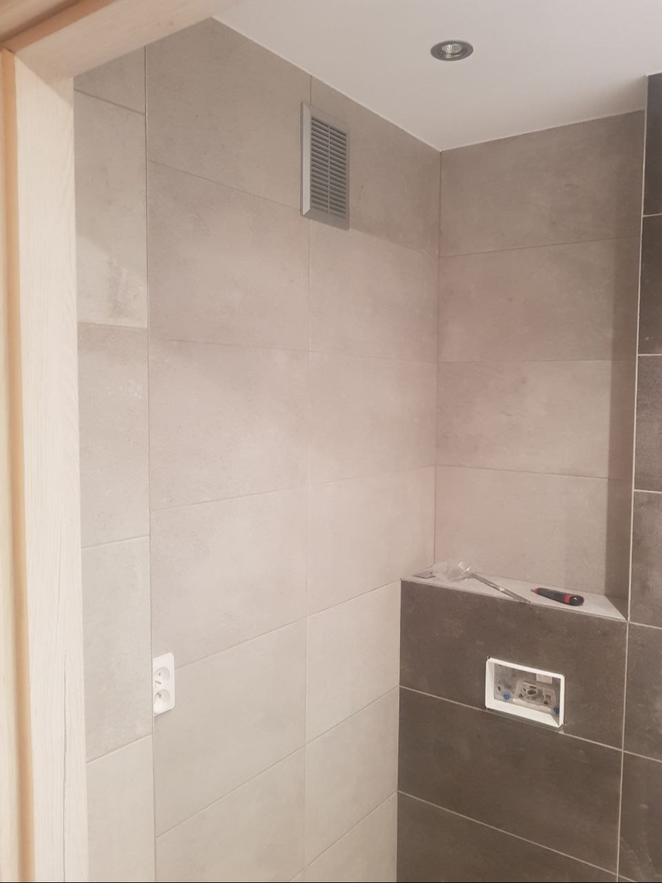 Wykończona łazienka w trakcie prac: jasne płytki na ścianach, ciemne płytki na półce, widoczna kratka wentylacyjna, gniazdko elektryczne i puszka pod włącznik, narzędzia na półce.