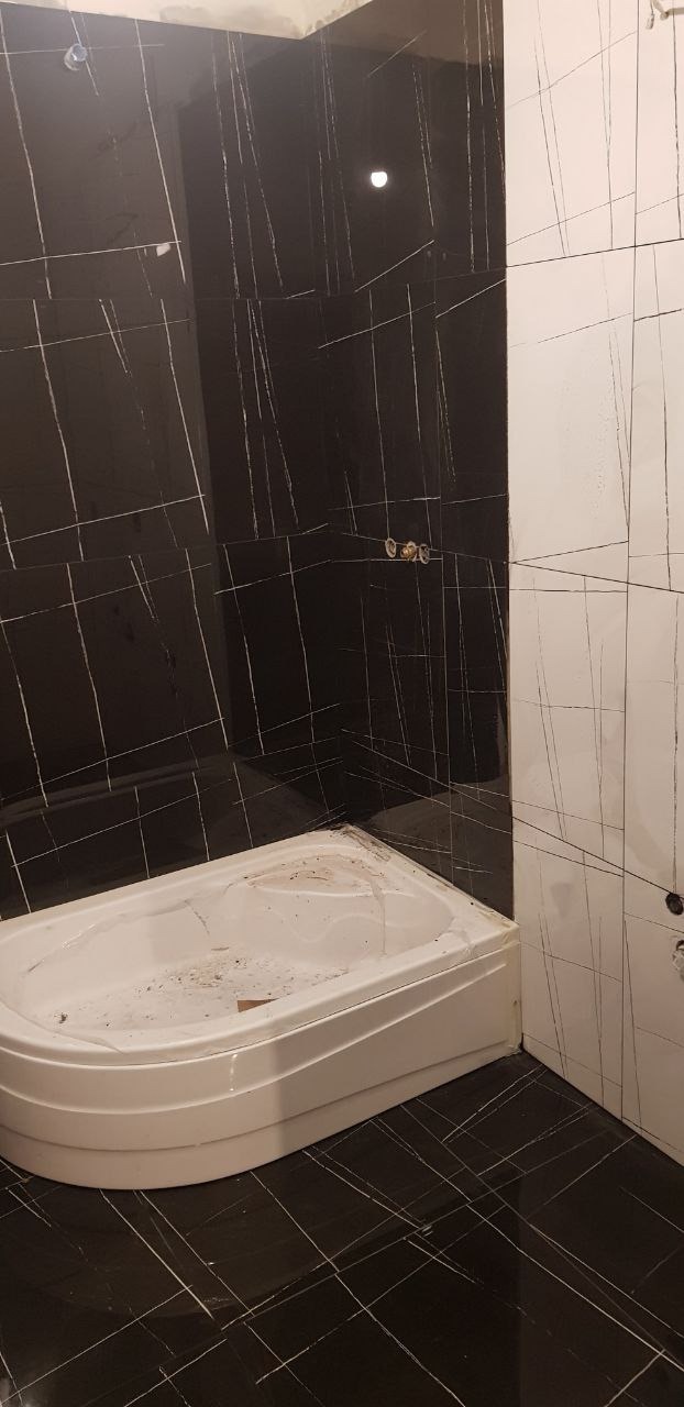 Niedokończona łazienka z czarnymi płytkami imitującymi marmur z białymi żyłami na ścianach i podłodze, biała wanna narożna w trakcie montażu, widoczne ślady po pracach wykończeniowych.