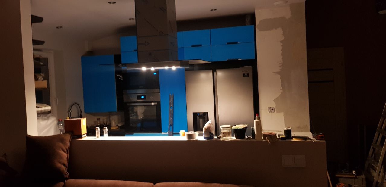 Niedokończona kuchnia z niebieskimi szafkami, widoczna ściana w trakcie przygotowania do wykończenia, narzędzia i materiały budowlane na blacie, w tle sprzęt AGD.