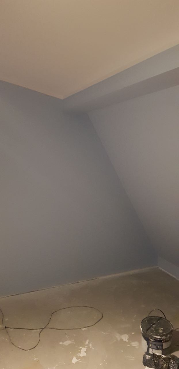 Pomalowane na szaro poddasze ze skośnym sufitem, widoczny kabel i wiadro po farbie na podłodze.