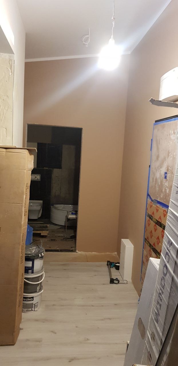 Wnętrze w trakcie remontu: świeżo pomalowane ściany w odcieniach beżu, widoczne puszki z farbą, w tle wykończona łazienka z białą armaturą, podłoga wyłożona panelami.