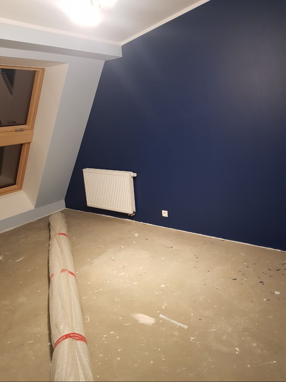 Pokój na poddaszu po malowaniu na granatowo, widoczny grzejnik, okno dachowe i rolka folii zabezpieczającej podłogę.