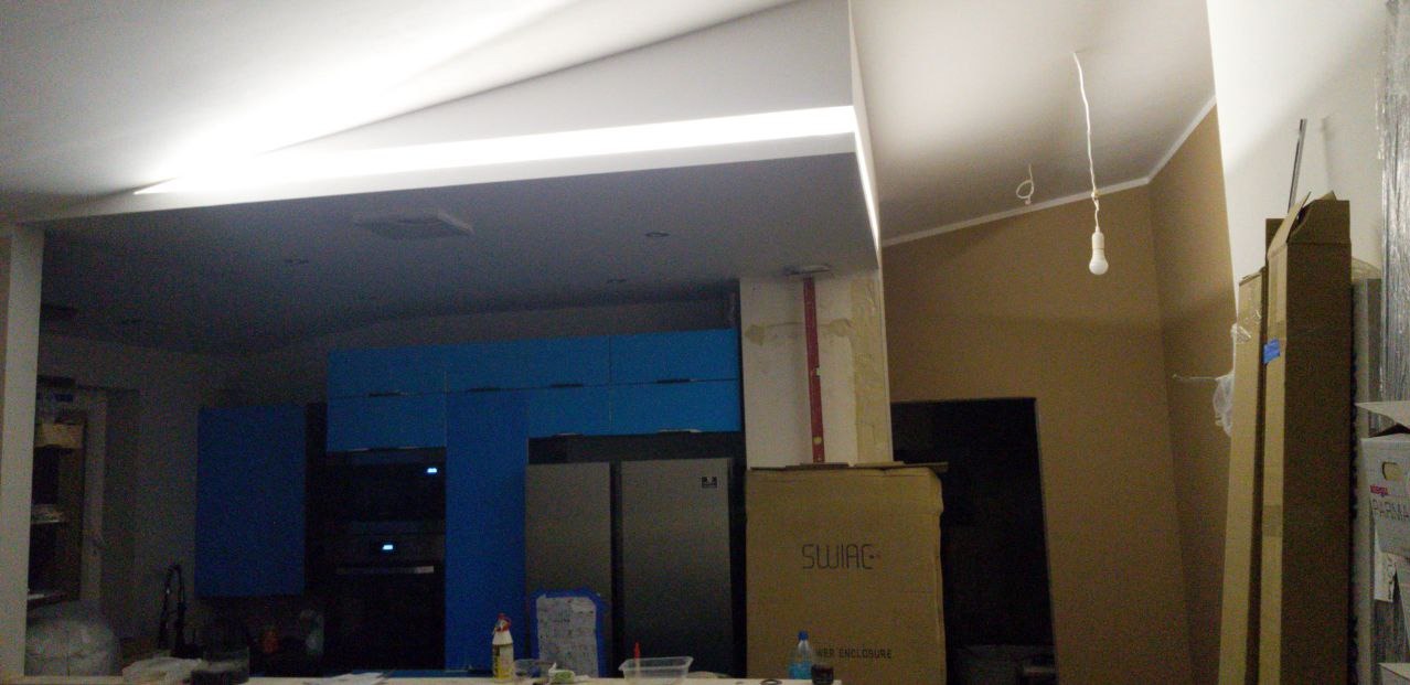 Wnętrze w trakcie prac wykończeniowych: widoczne niebieskie szafki kuchenne, surowe ściany przygotowane do malowania, wystająca żarówka na kablu i kartony z materiałami budowlanymi.