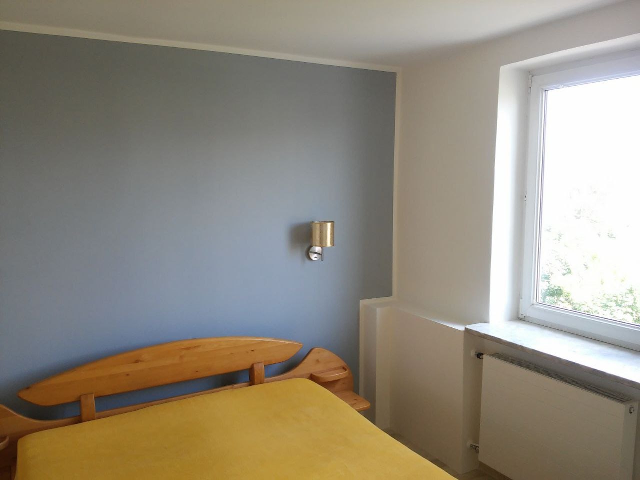 Pomieszczenie po malowaniu ścian na szaro, z drewnianym łóżkiem, lampką nocną i białym oknem z widokiem na zieleń.