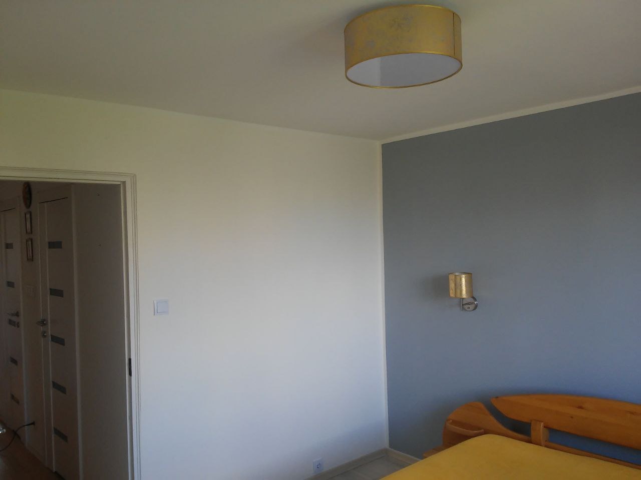 Pokój po odświeżeniu: biała ściana z włącznikiem światła sąsiaduje z szarą ścianą, na której wisi złoty kinkiet. Widoczna część drewnianego łóżka z żółtą narzutą i lampa sufitowa z abażurem...