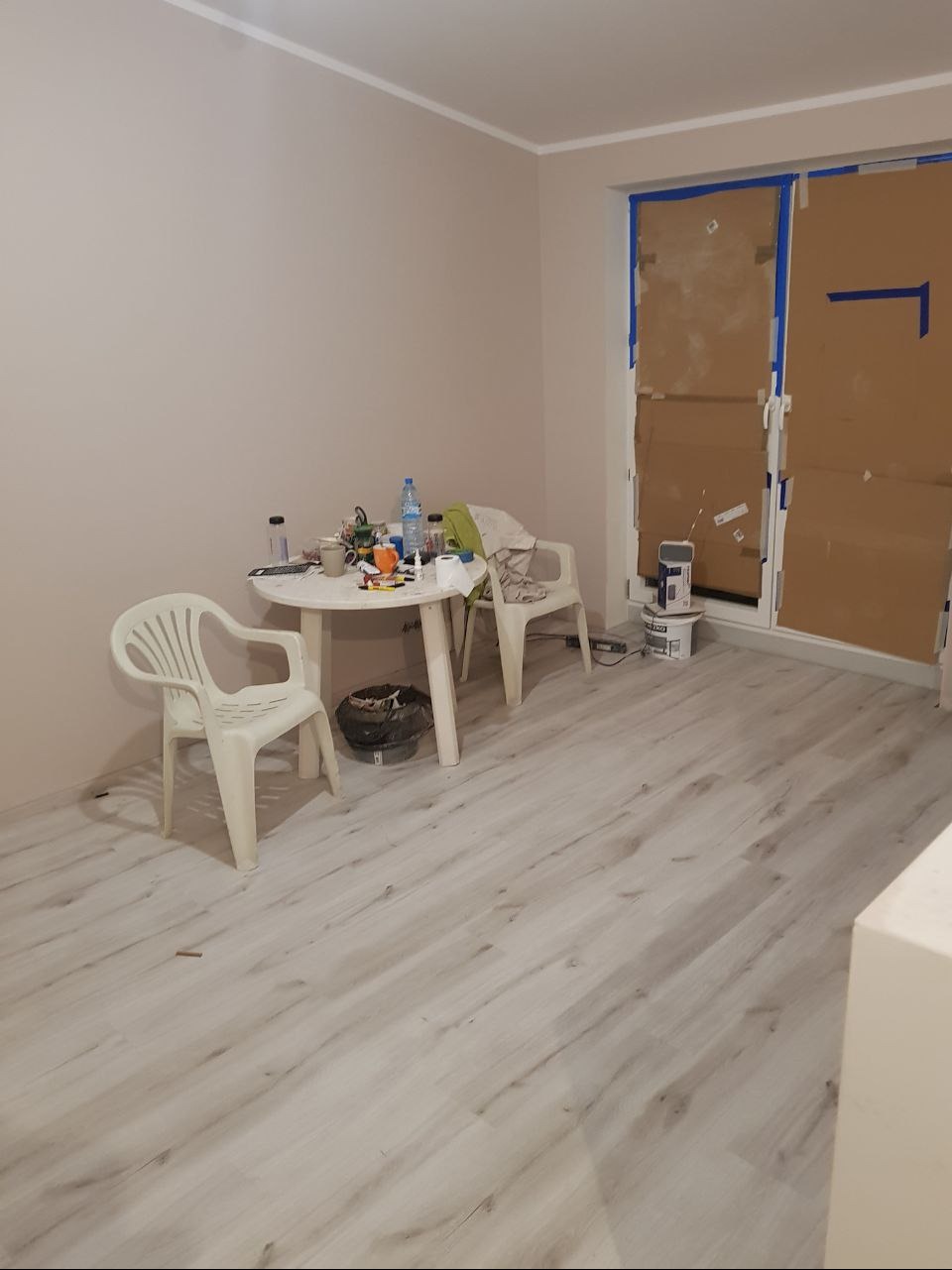 Pokój w trakcie odświeżania: ściany świeżo pomalowane na jasny beż, okno zabezpieczone kartonem i niebieską taśmą malarską, plastikowe krzesła i okrągły stolik z akcesoriami malarskimi w centrum.