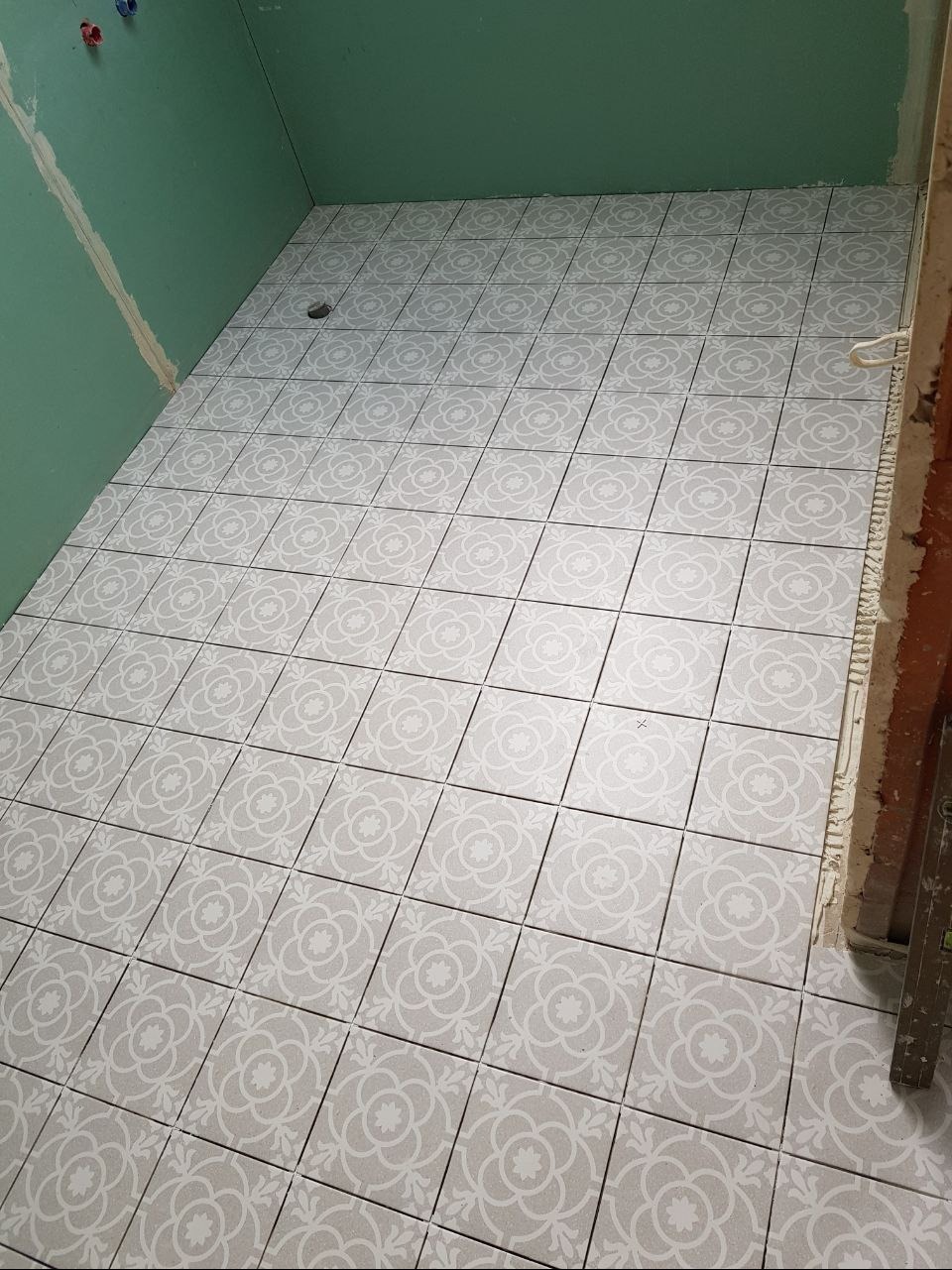 Ułożone szare płytki podłogowe z białym kwiatowym wzorem w łazience w trakcie remontu, widoczne niezakończone ściany z płyt kartonowo-gipsowych.