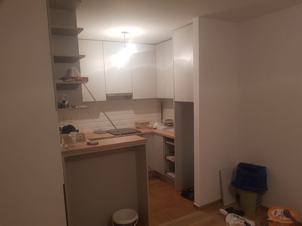 Wnętrze kuchni w trakcie prac wykończeniowych: białe szafki kuchenne, białe płytki na ścianie, widoczne narzędzia i wiadro z farbą na podłodze, pomieszczenie w trakcie odświeżania.