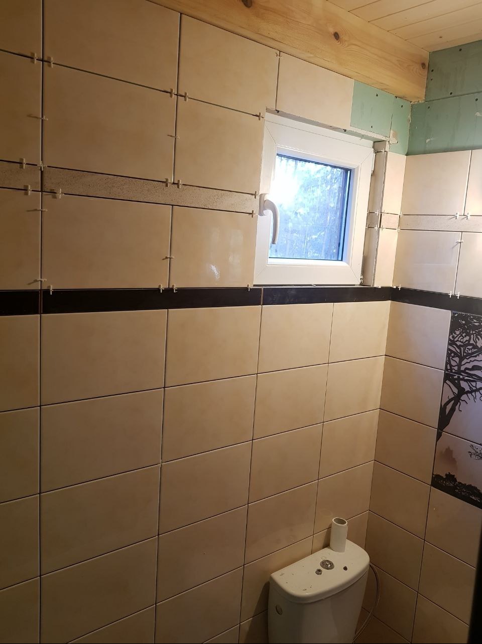 Beżowe płytki ścienne z czarnym pasem dekoracyjnym w trakcie układania w łazience, widoczne krzyżyki dystansowe, okno z widokiem na zewnątrz i fragment sufitu z drewnianą belką.