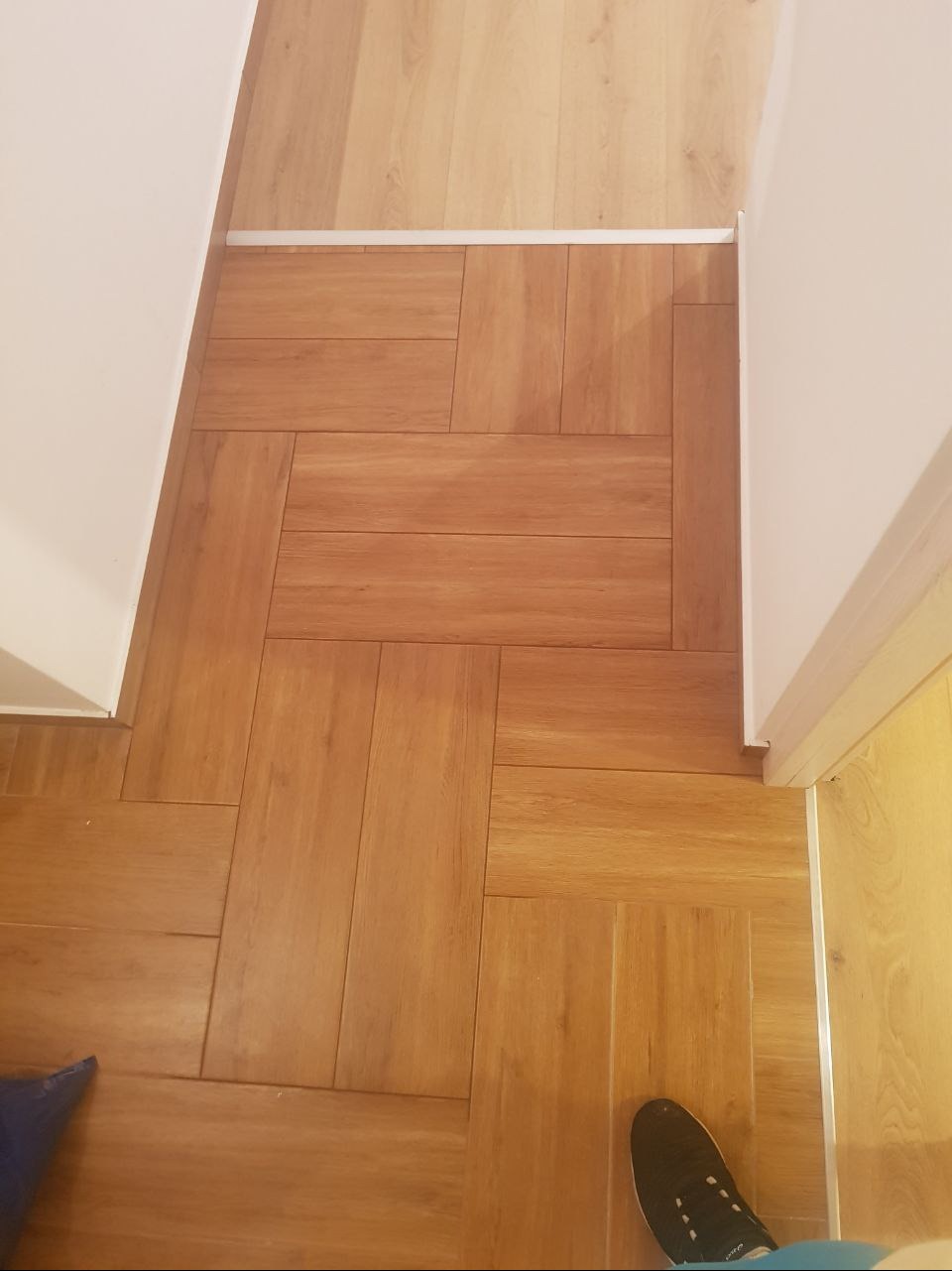 Ułożona parkietowa podłoga w odcieniach brązu, z widocznym fragmentem buta i niebieską torbą w rogu, przy białej ścianie i futrynie drzwi.