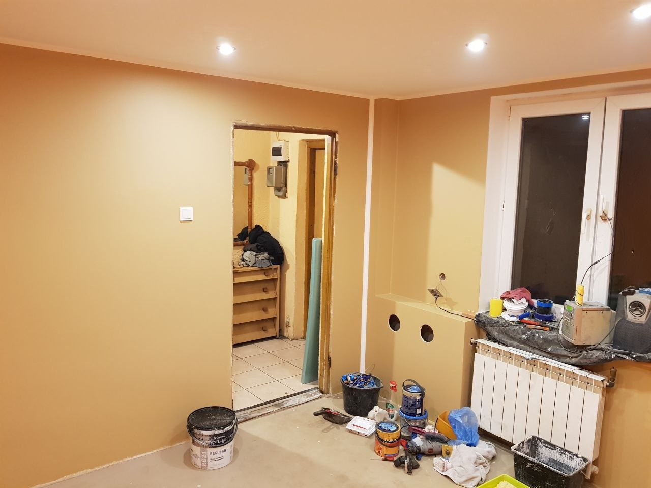 Pokój w trakcie odświeżania: ściany pomalowane na beżowo, widoczne puszki z farbą, narzędzia malarskie i grzejnik pod oknem. W tle otwarte drzwi do innego pomieszczenia z regałem.