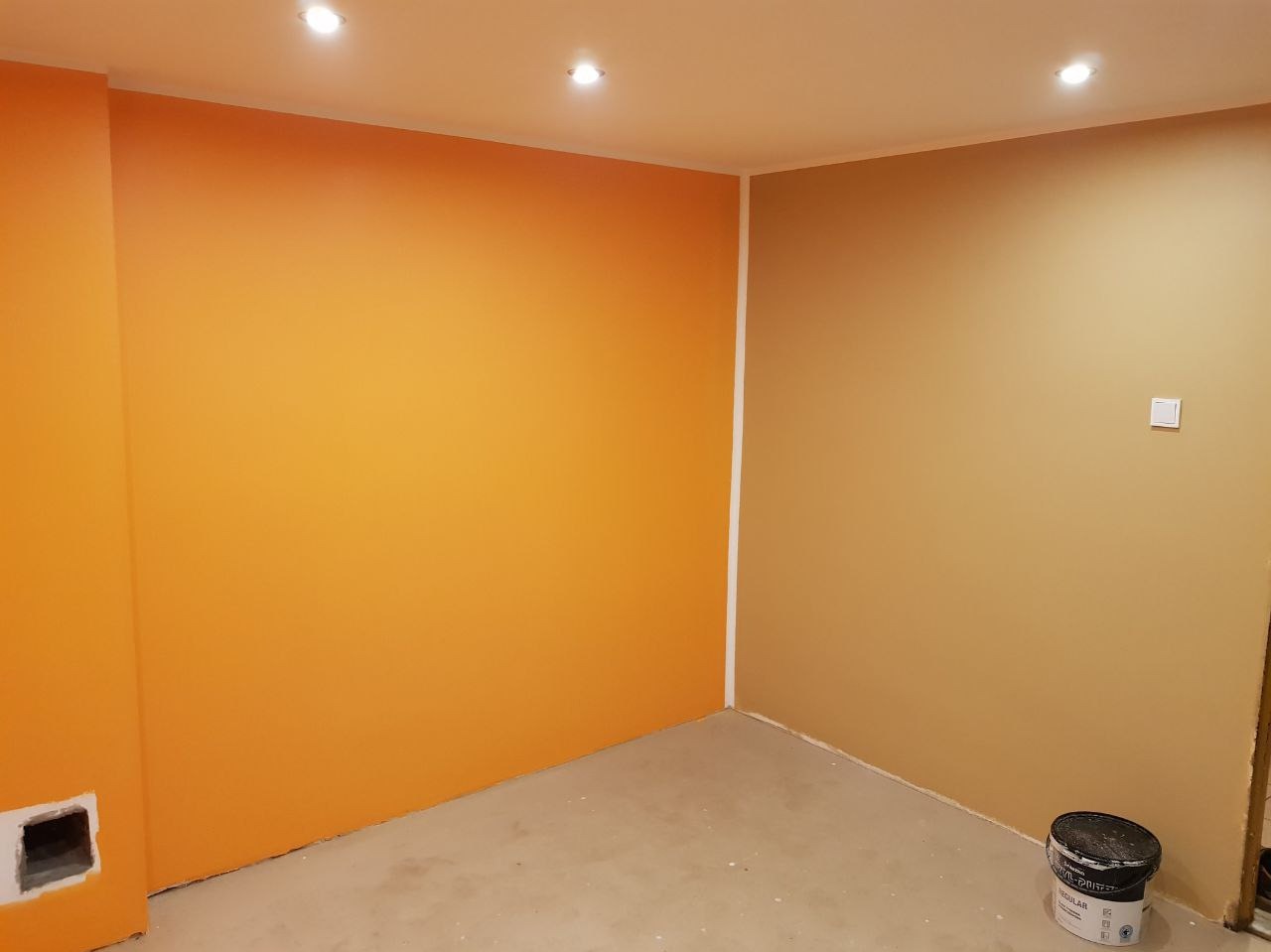 Pomarańczowa i beżowa ściana w pokoju po świeżym pomalowaniu, widoczna puszka z farbą przy podłodze.