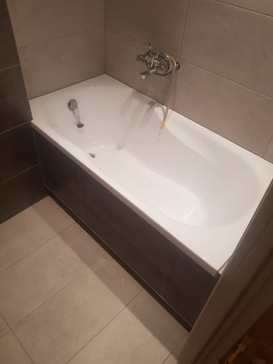 Wanna z obudową z ciemnych paneli, chromowana bateria i odpływ, beżowe płytki na ścianach i podłodze, widok z góry pod kątem.