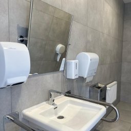 PAKETPRO SPÓŁKA Z OGRANICZONĄ ODPOWIEDZIALNOŚCIĄ - Umywalka z chromowanymi uchwytami dla niepełnosprawnych, szare płytki, lustro, dozownik mydła i suszarka do rąk w nowoczesnej łazience publicznej.