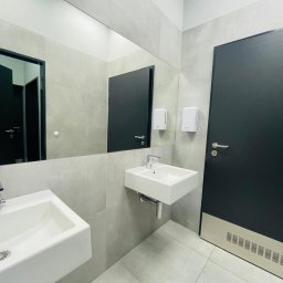 PAKETPRO SPÓŁKA Z OGRANICZONĄ ODPOWIEDZIALNOŚCIĄ - Nowoczesna łazienka w szarościach: dwie białe umywalki, duże lustro, czarne drzwi. Minimalistyczny design z dozownikami mydła na ścianie i jasną podłogą.