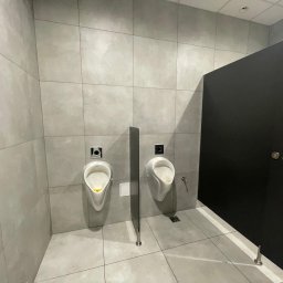 PAKETPRO SPÓŁKA Z OGRANICZONĄ ODPOWIEDZIALNOŚCIĄ - Wykończona toaleta publiczna: dwa białe pisuary na szarej ścianie z płytek, oddzielone pionową przegrodą. Czarne drzwi kabiny po prawej stronie. Minimalistyczny design.