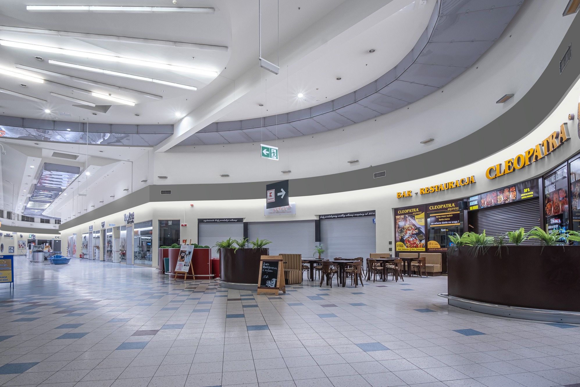 Wnętrze galerii handlowej z zamkniętymi lokalami i stolikami restauracyjnymi, widok na kafelkowaną podłogę i sufit z oświetleniem.