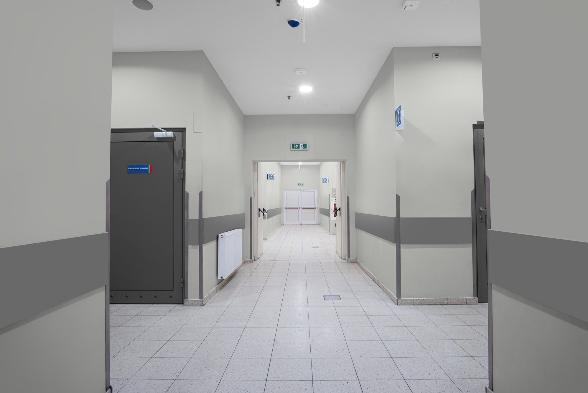Korytarz w jasnej tonacji z szarymi odbojami ściennymi i płytkami podłogowymi. Widoczne drzwi techniczne i ewakuacyjne, oświetlenie sufitowe i znaki informacyjne.