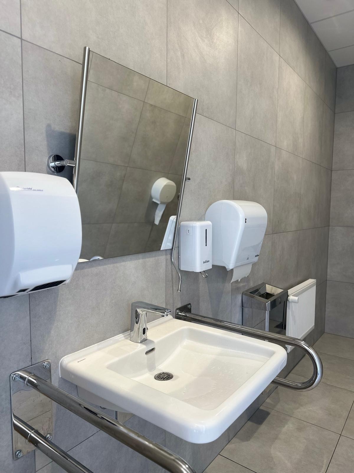 Umywalka z chromowanymi uchwytami dla niepełnosprawnych, szare płytki, lustro, dozownik mydła i suszarka do rąk w nowoczesnej łazience publicznej.