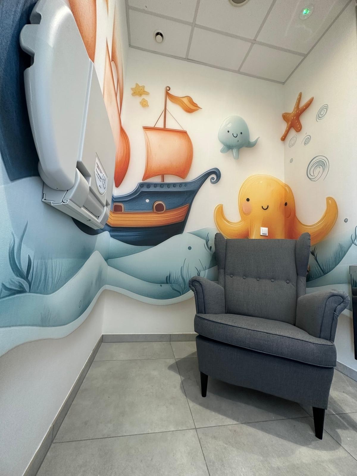 Pokój dziecięcy z marynistycznym muralem: statek, ośmiornica, gwiazdy. Szary fotel klubowy na tle ściany z rysunkami. Na ścianie przewijak.