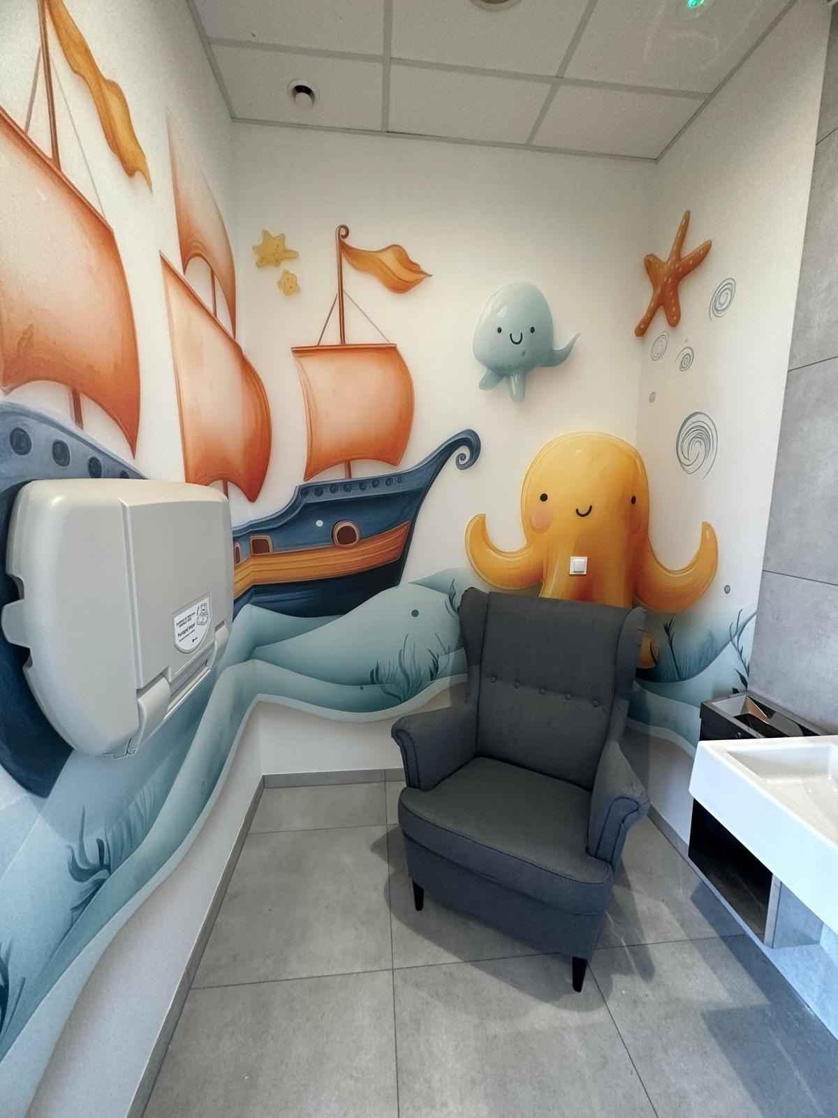 Pokój dziecięcy z marynistycznym muralem: okręt, ośmiornica, fotel uszak i przewijak. Aranżacja wnętrza z akcentem morskim, w jasnej tonacji.
