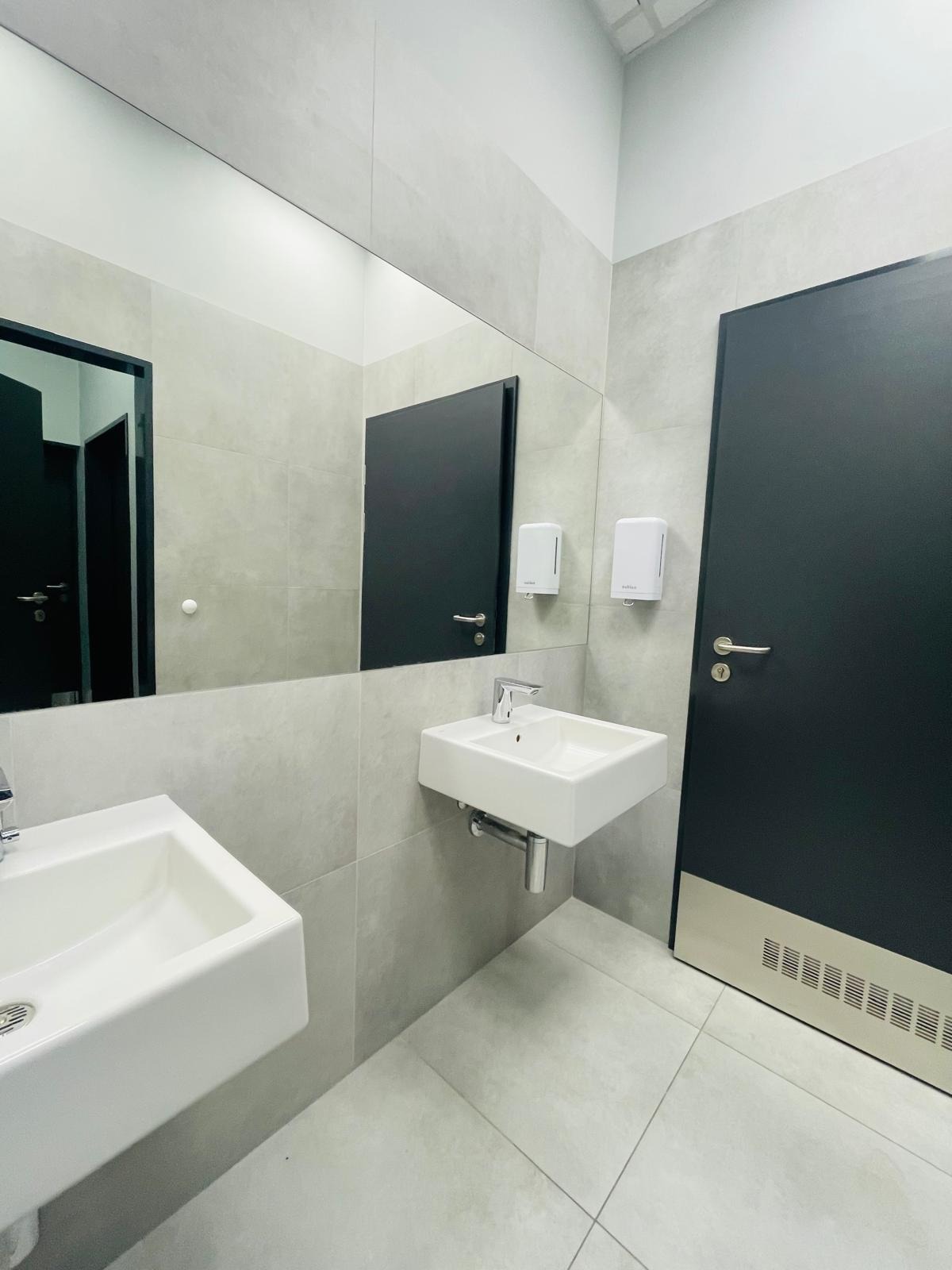 Nowoczesna łazienka w szarościach: dwie białe umywalki, duże lustro, czarne drzwi. Minimalistyczny design z dozownikami mydła na ścianie i jasną podłogą.