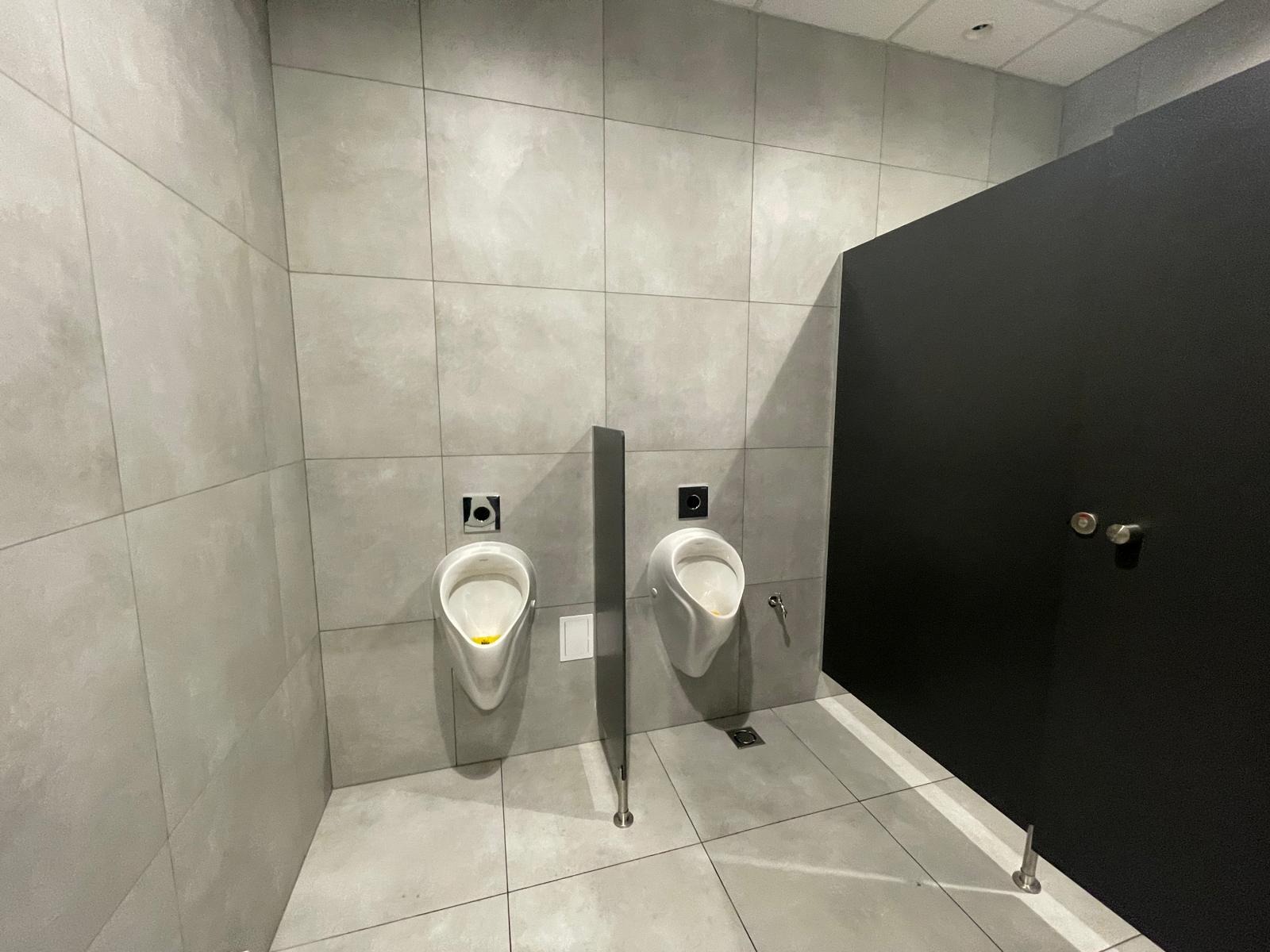 Wykończona toaleta publiczna: dwa białe pisuary na szarej ścianie z płytek, oddzielone pionową przegrodą. Czarne drzwi kabiny po prawej stronie. Minimalistyczny design.