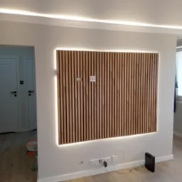 Drewniana ściana dekoracyjna z podświetleniem LED, gniazda elektryczne i puszka instalacyjna, pomalowane ściany na biało, widoczne drzwi i podłoga.