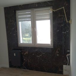 Gres bud - Wykończona ściana z ciemnego marmuru wokół okna z roletami, z zamontowanym grzejnikiem i złotą lampą ścienną.