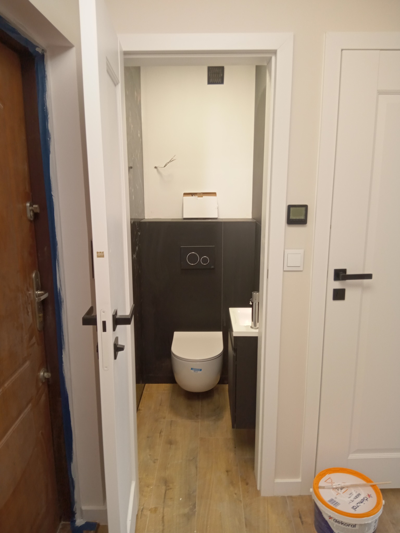 Wnętrze wykończonej łazienki z toaletą, umywalką i czarnymi płytkami częściowo wyłożonymi na ścianie, widoczne zza otwartych drzwi, obok których stoi wiadro z farbą.