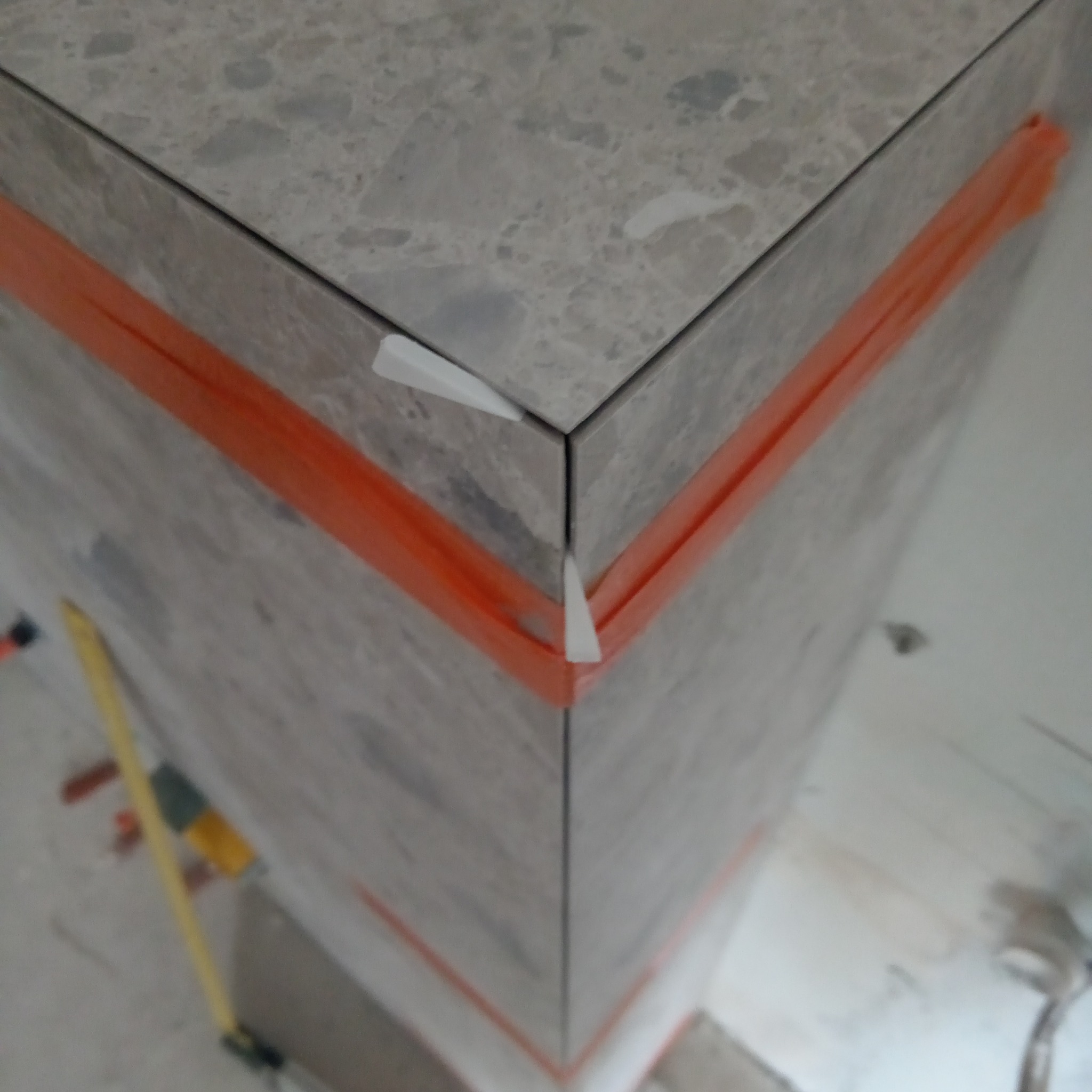 Fragment narożnika obłożonego marmurowymi płytkami, zabezpieczonego pomarańczową taśmą i białymi krzyżykami dystansowymi, widoczny poziomica w tle.
