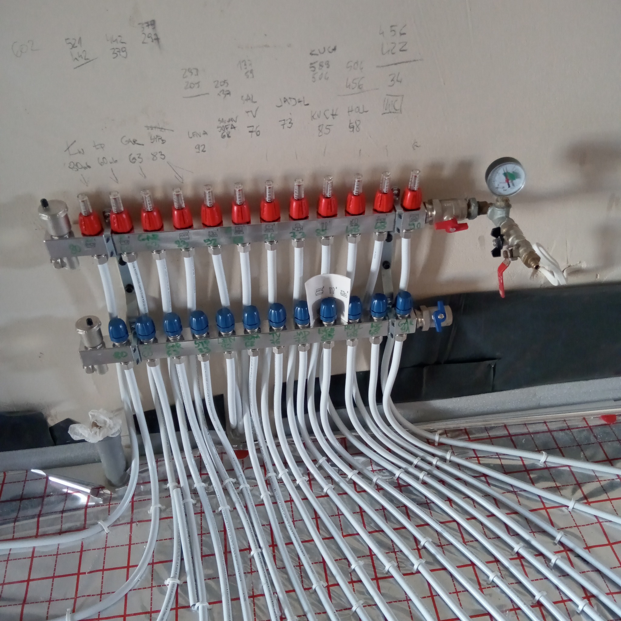Rozdzielacz ogrzewania podłogowego z białymi rurkami na folii izolacyjnej z czerwoną kratką, zamontowany na ścianie z odręcznymi notatkami i numerami.