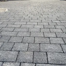 Szara kostka brukowa ułożona w regularny wz&oacute;r, z widocznym włazem kanalizacyjnym w lewym g&oacute;rnym rogu i fragmentem trawnika w tle. Widok z perspektywy.