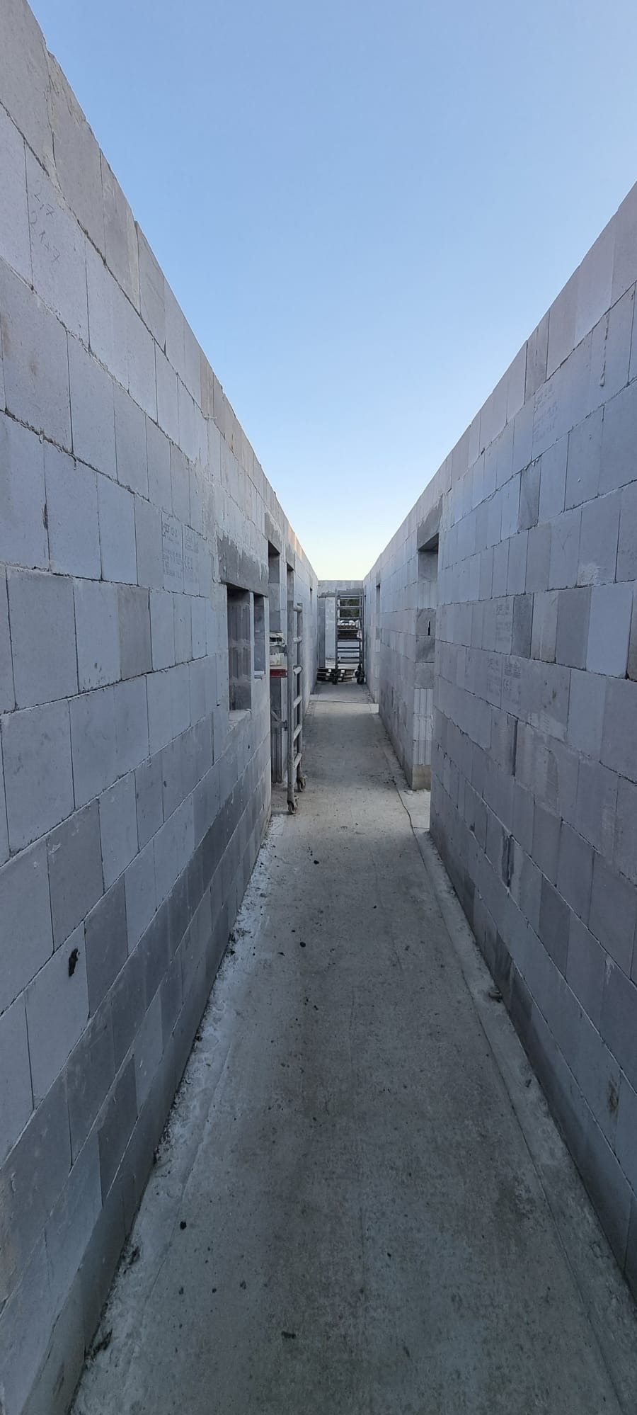 Wnętrze budynku w trakcie budowy, widoczne ściany z szarych bloczków, otwory okienne i betonowa posadzka. W tle widoczne rusztowania i materiały budowlane.