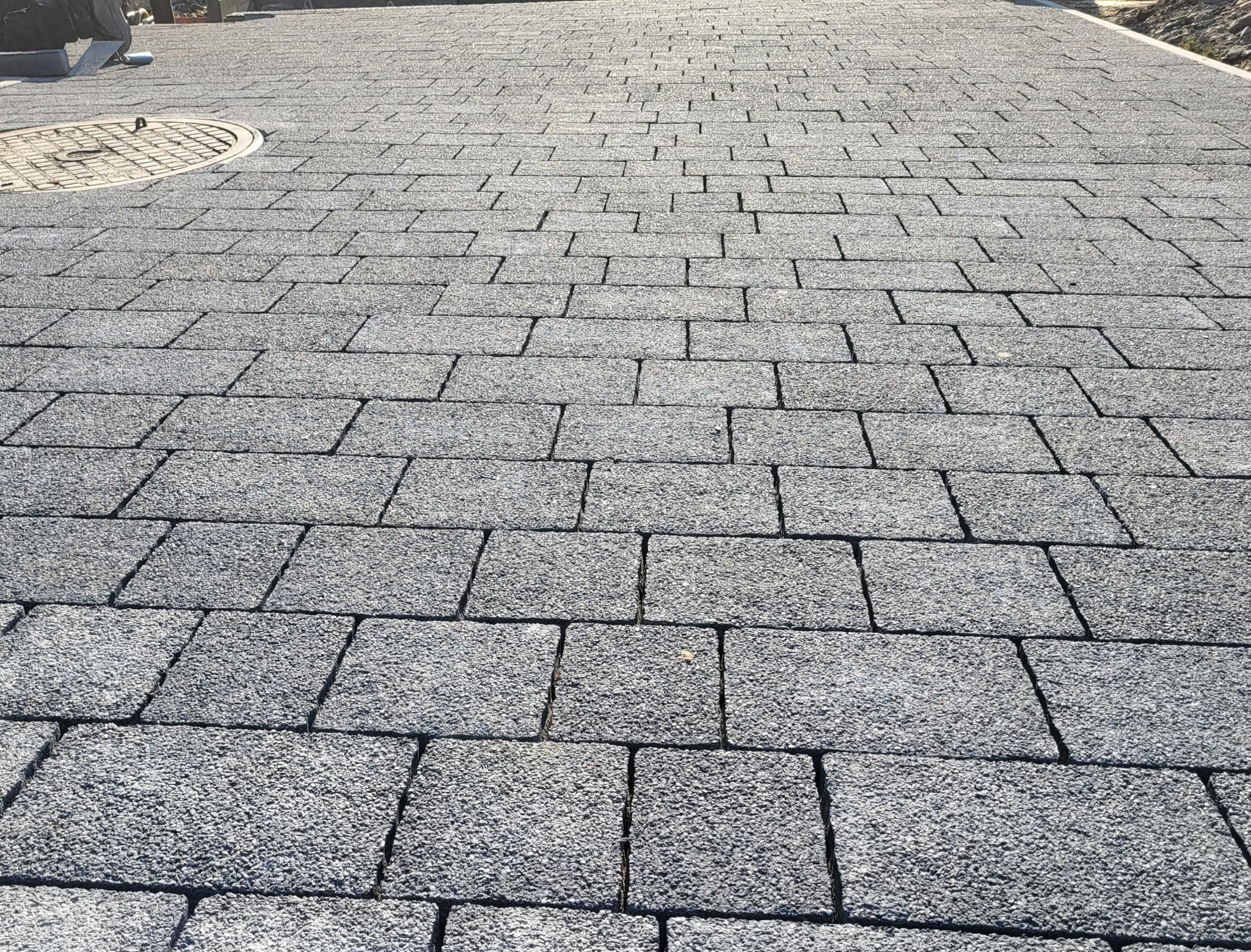 Szara kostka brukowa ułożona w regularny wzór, z widocznym włazem kanalizacyjnym w lewym górnym rogu i fragmentem trawnika w tle. Widok z perspektywy.