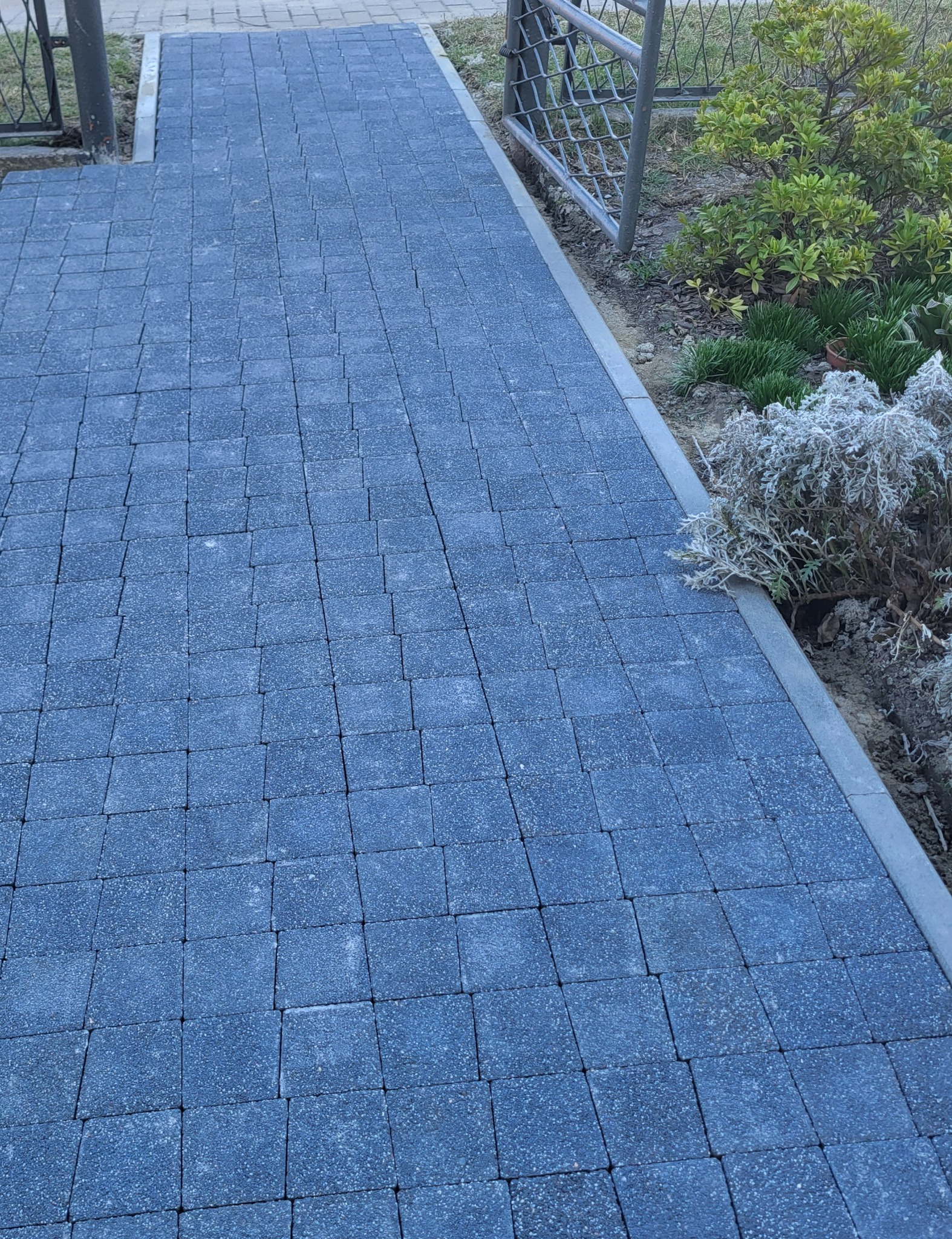Ułożona, grafitowa kostka brukowa tworząca ścieżkę w ogrodzie, z widocznym obrzeżem i fragmentem metalowej furtki. Obok rosną krzewy i trawa.