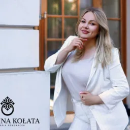 Portret uśmiechniętej kobiety w białym garniturze, opierającej się o biały narożnik budynku z widocznym logo kancelarii adwokackiej w lewym dolnym rogu.