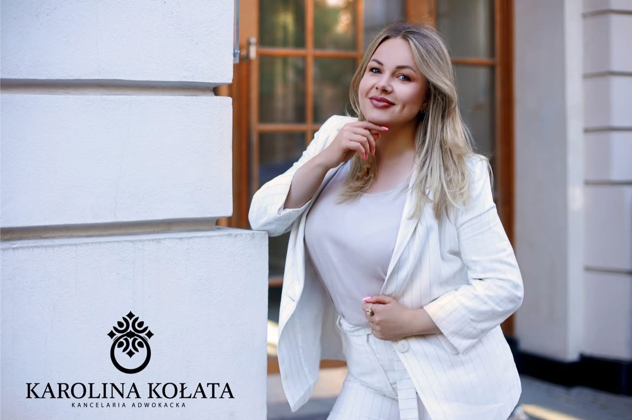 Portret uśmiechniętej kobiety w białym garniturze, opierającej się o biały narożnik budynku z widocznym logo kancelarii adwokackiej w lewym dolnym rogu.