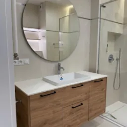 Nowoczesna łazienka z szafką pod umywalką z drewnopodobnymi frontami, białym blatem i okrągłym lustrem, kabina prysznicowa w tle.