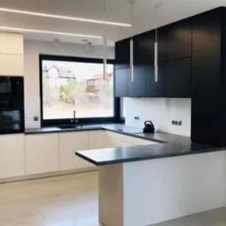 Nowoczesna kuchnia w bieli i czerni z wyspą, oświetleniem LED i dużym oknem z widokiem na ogród. Minimalistyczny design z elementami dekoracyjnymi.