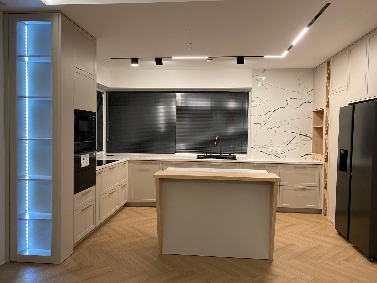 Nowoczesna, beżowa kuchnia z wyspą i podświetlaną witryną, parkiet w jodełkę, marmurowy backsplash i czarne akcenty oświetleniowe.