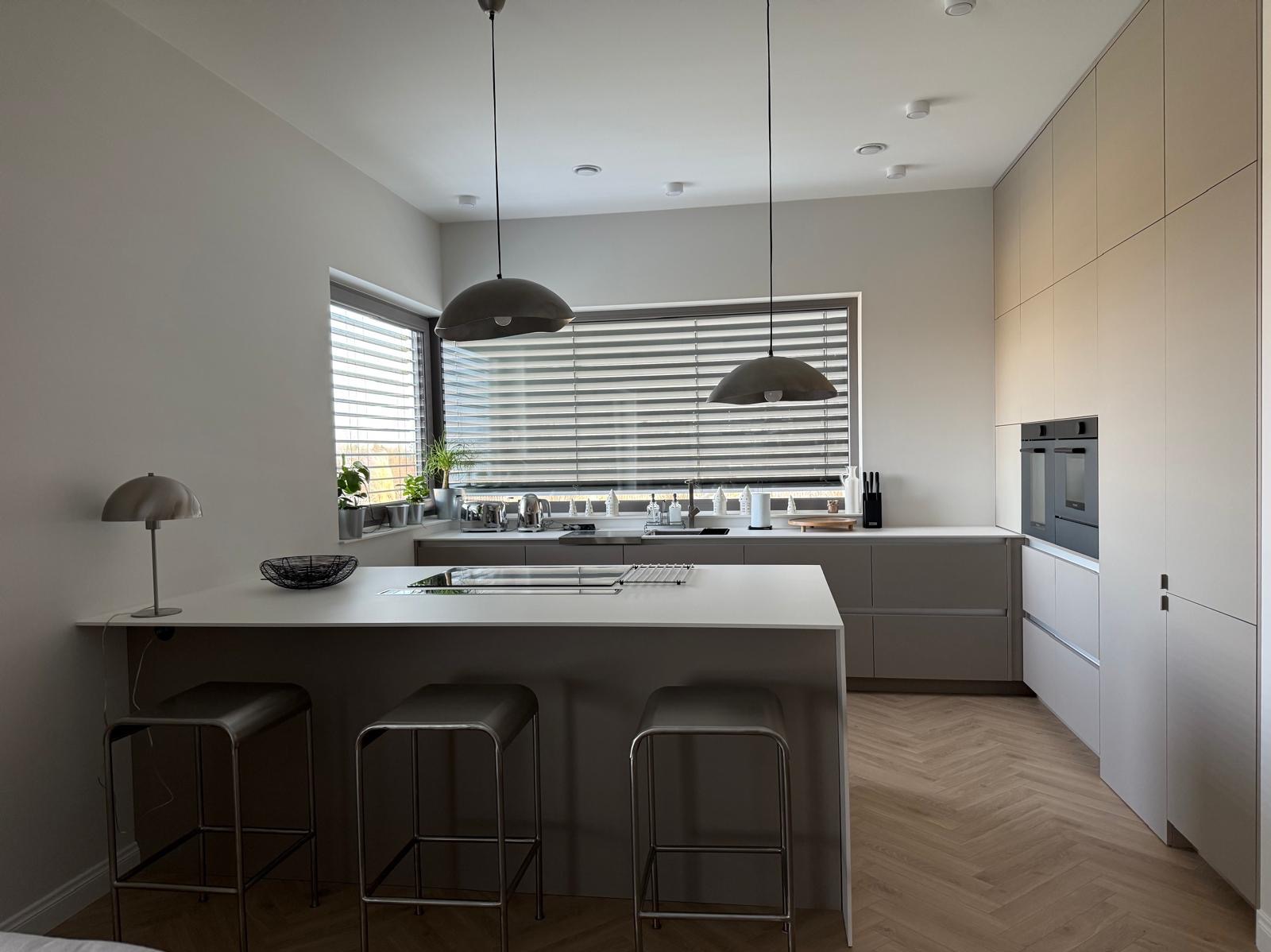 Jasna, nowoczesna kuchnia z wyspą, trzema hokerami, dwiema lampami wiszącymi i parkietem w jodełkę; widok na okno z żaluzjami.