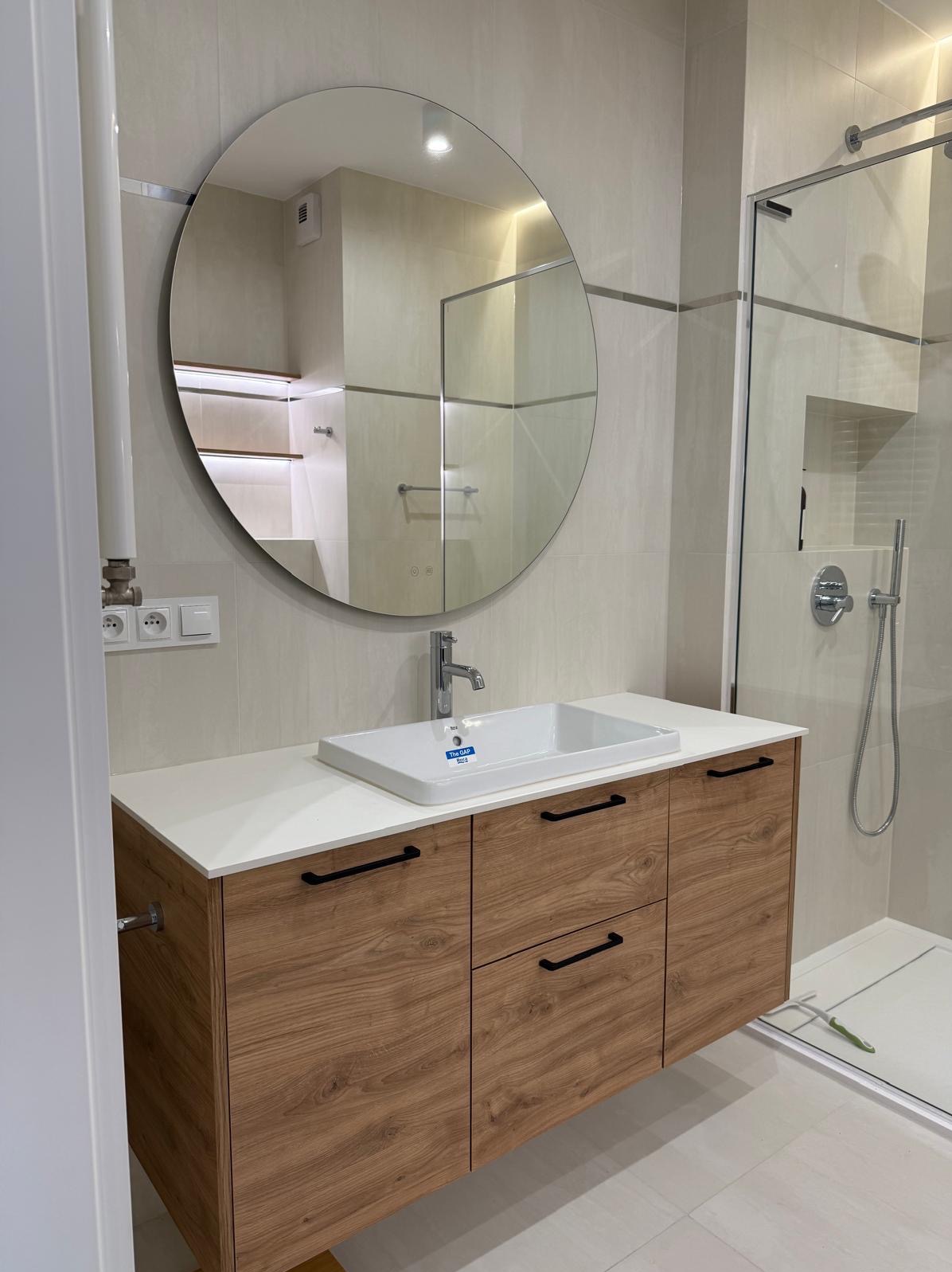 Nowoczesna łazienka z szafką pod umywalką z drewnopodobnymi frontami, białym blatem i okrągłym lustrem, kabina prysznicowa w tle.
