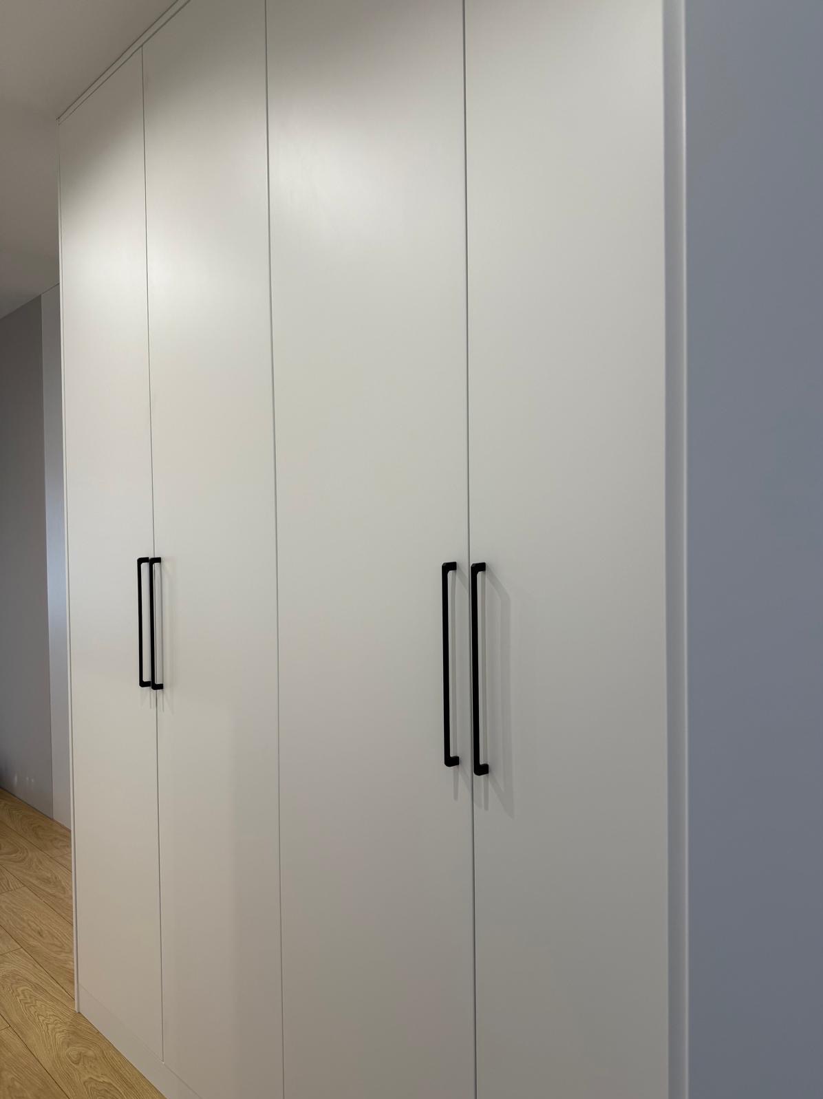 Biała, minimalistyczna szafa wnękowa z czarnymi, pionowymi uchwytami na tle jasnoszarej ściany i drewnianej podłogi.