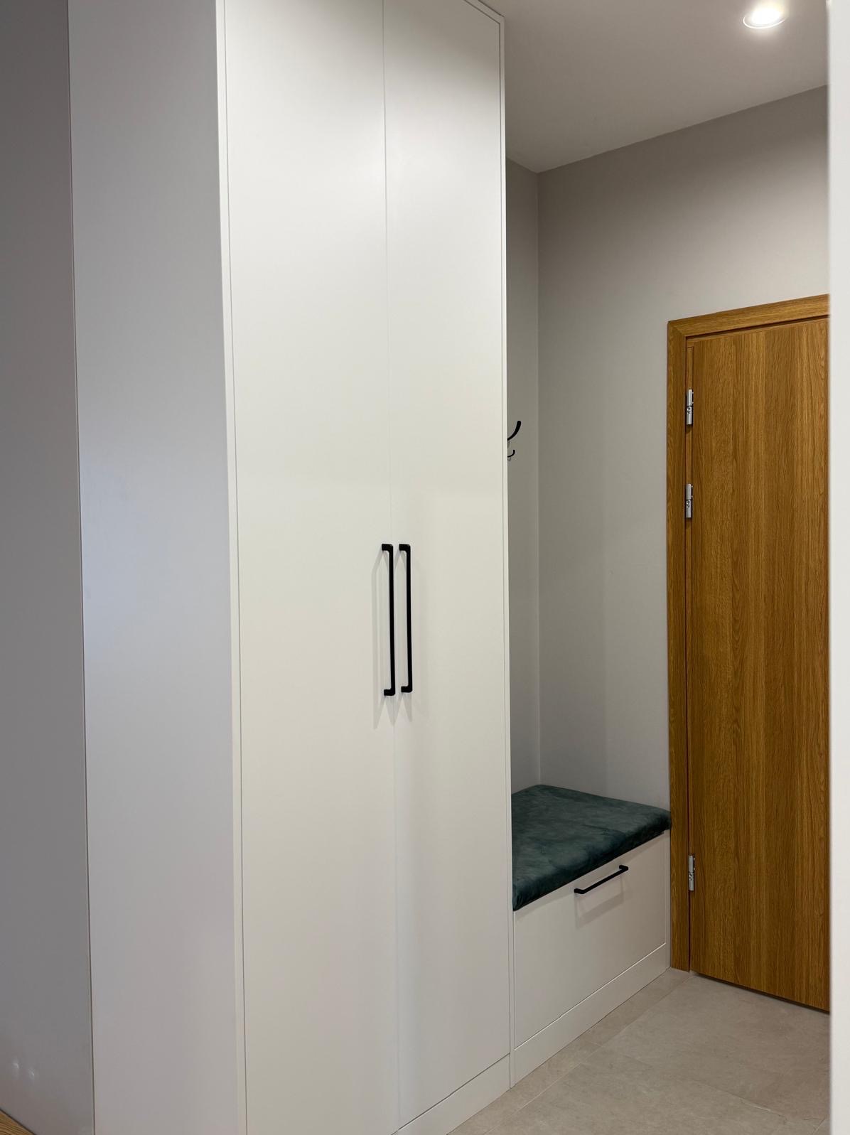 Biała szafa wnękowa z czarnymi uchwytami, połączona z siedziskiem z zieloną tapicerką w przedpokoju, obok drewniane drzwi.